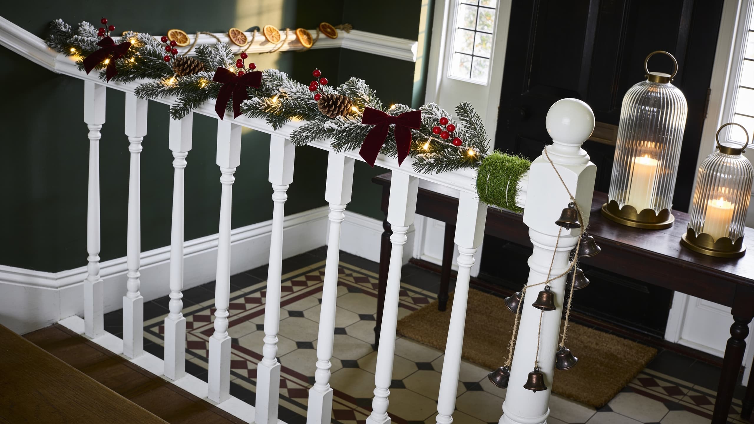 A Victorian terrace hallway at Christmas, dark green walls and white woodwork: a snow-tipped pre-lit pine garland with burgundy velvet bows up the banister, held by a green grass-covered tie, a jute rope of bronze bells round the newel post, a dried orange garland along the dado rail, two ribbed glass lanterns with flameless candles on the hall table and a coir mat on the tiles