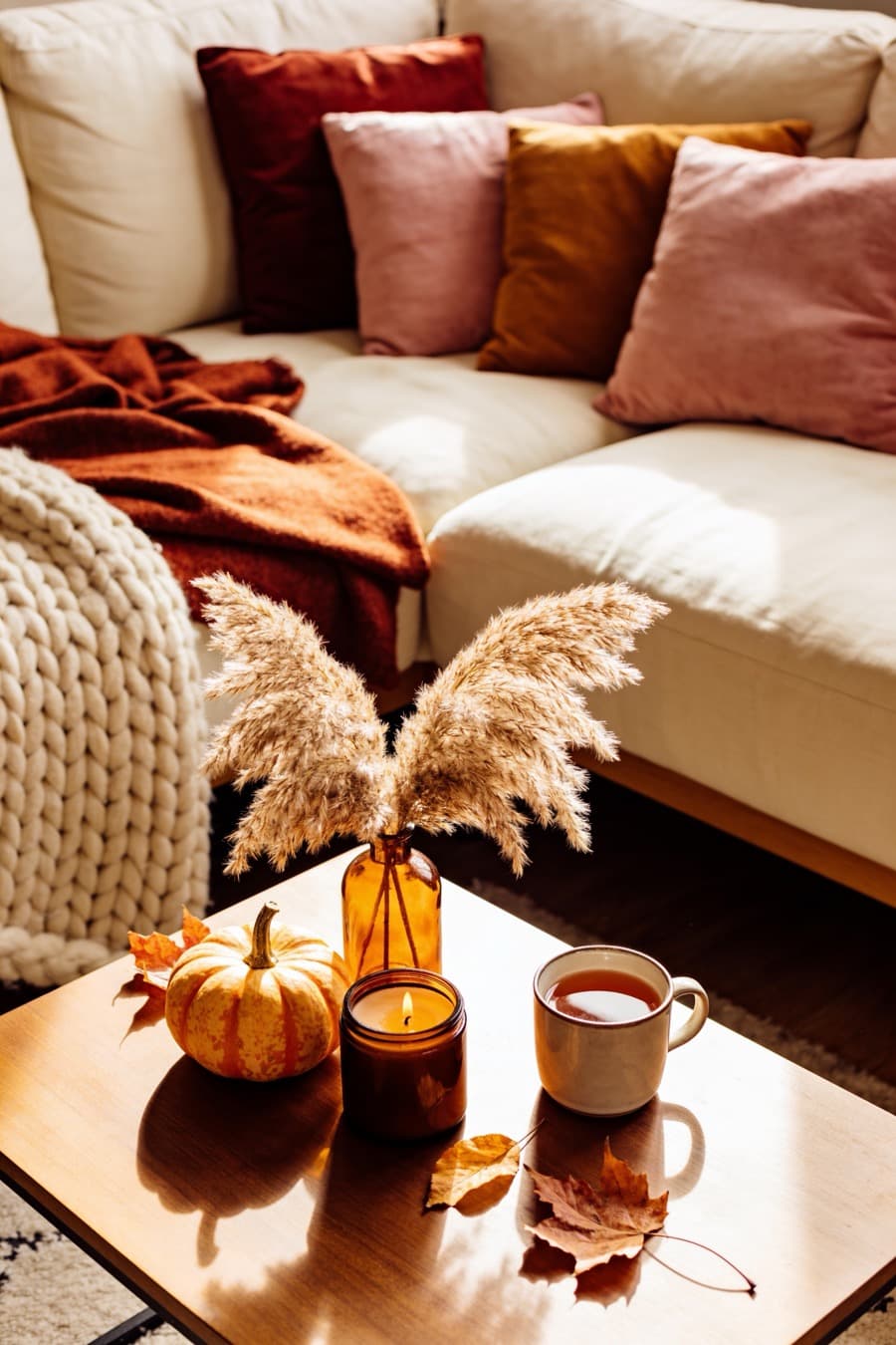 A cream sofa piled with rust and pink cushions and a chunky knit throw, a candle and a mug of tea on the coffee table