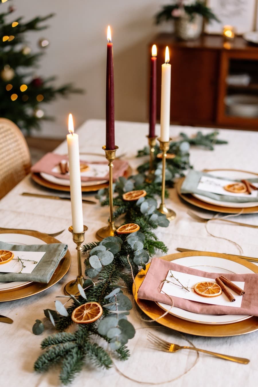 A Christmas table laid with a linen cloth, brass candlesticks, taper candles, eucalyptus and dried orange slices