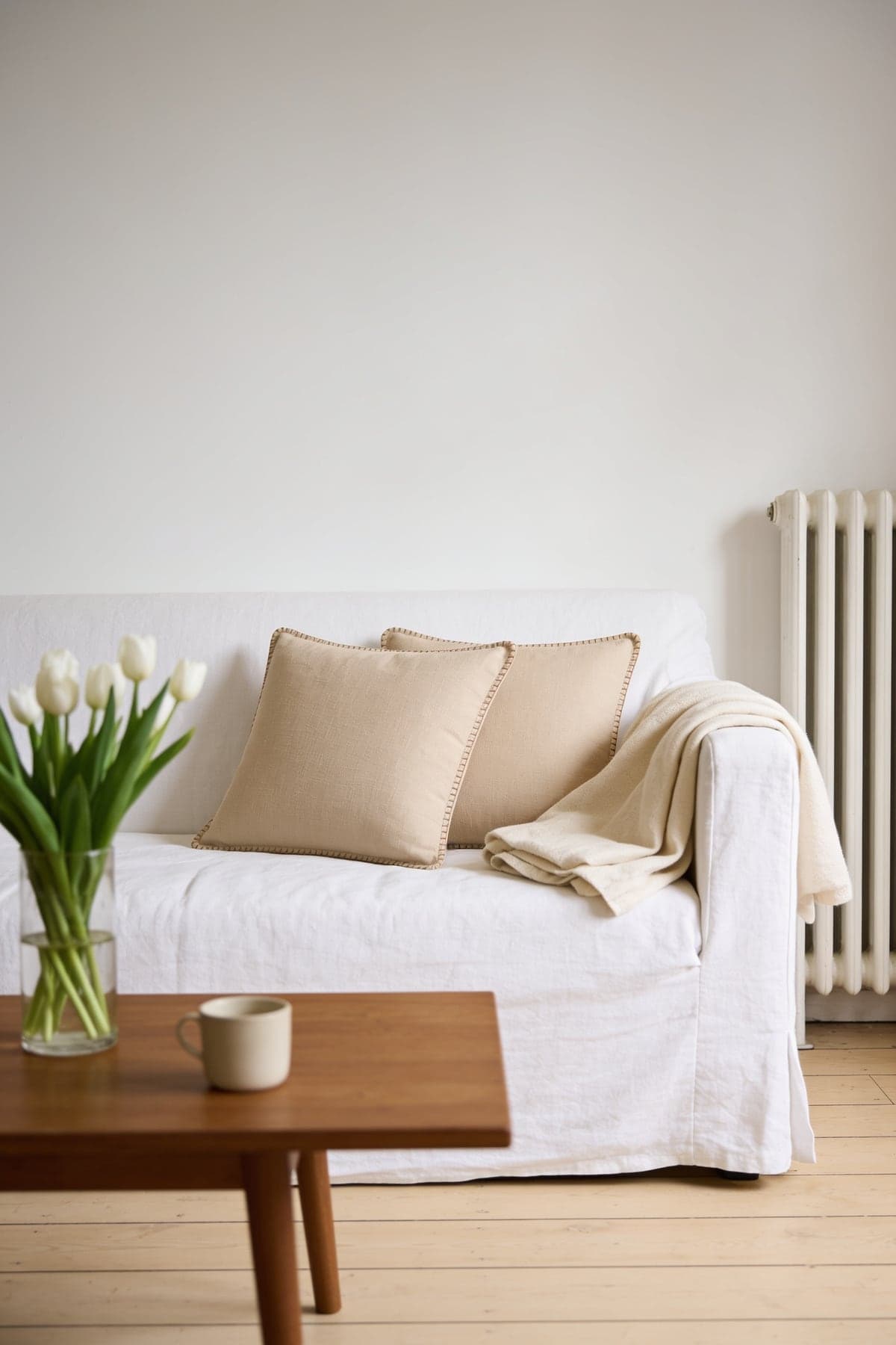 Two beige linen cushions with stitched edges on a white sofa beside tulips and a coffee table