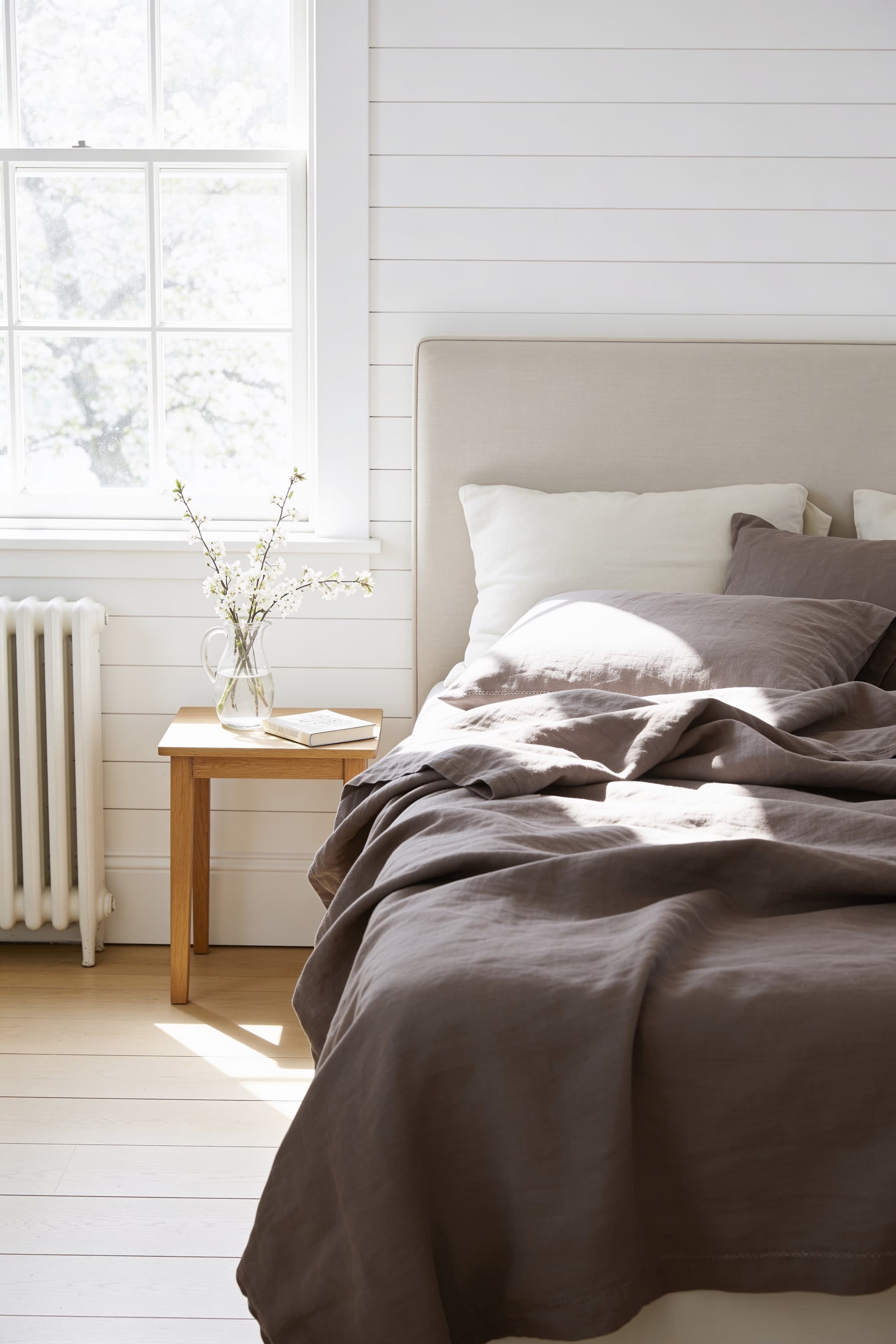 A bed made with taupe stonewashed linen bedding beside a sunny sash window, a small table with blossom in a jug