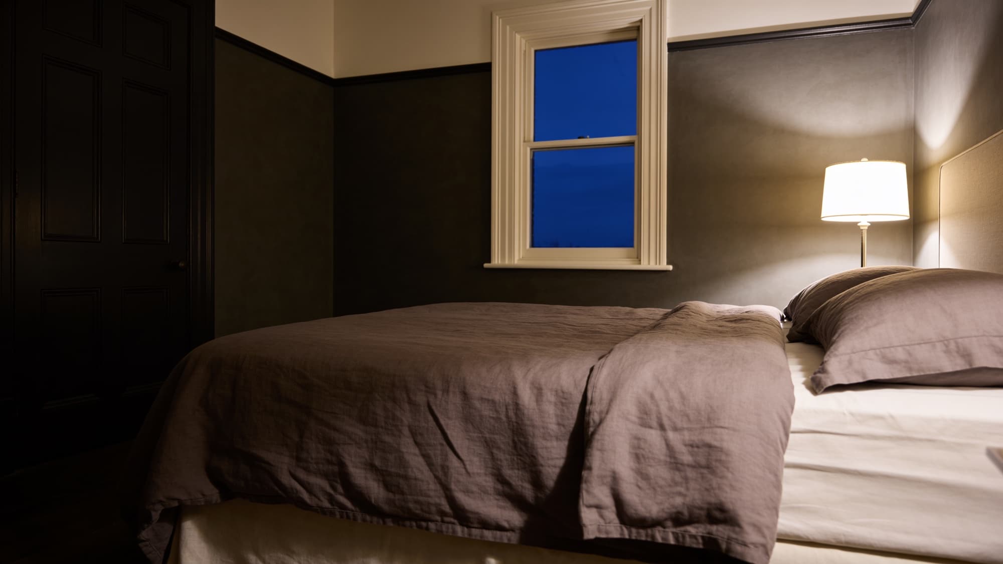A bed made in taupe stonewashed linen beside a lamp at dusk