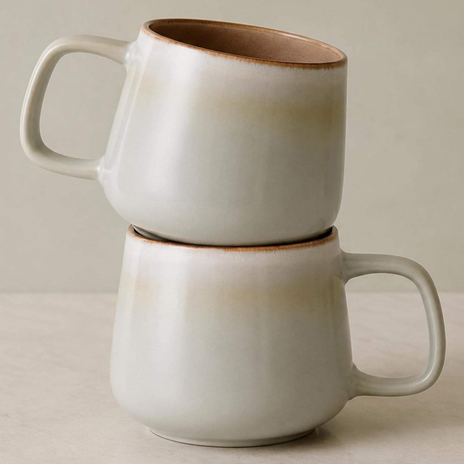 Two large sage-green stoneware mugs with a mottled reactive glaze on a pale wooden table, one holding a milky coffee