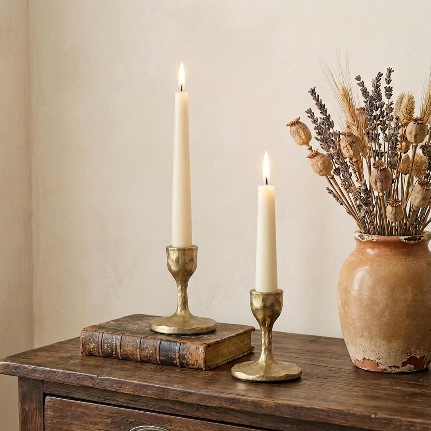 Two short hammered cast iron candle holders in an antique gold finish, each holding a cream taper, on a side table