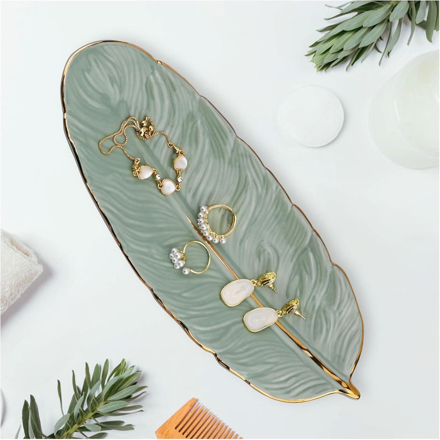 A glazed green ceramic dish shaped like a feather with gold-painted edges, holding two rings and a chain on a bedside table