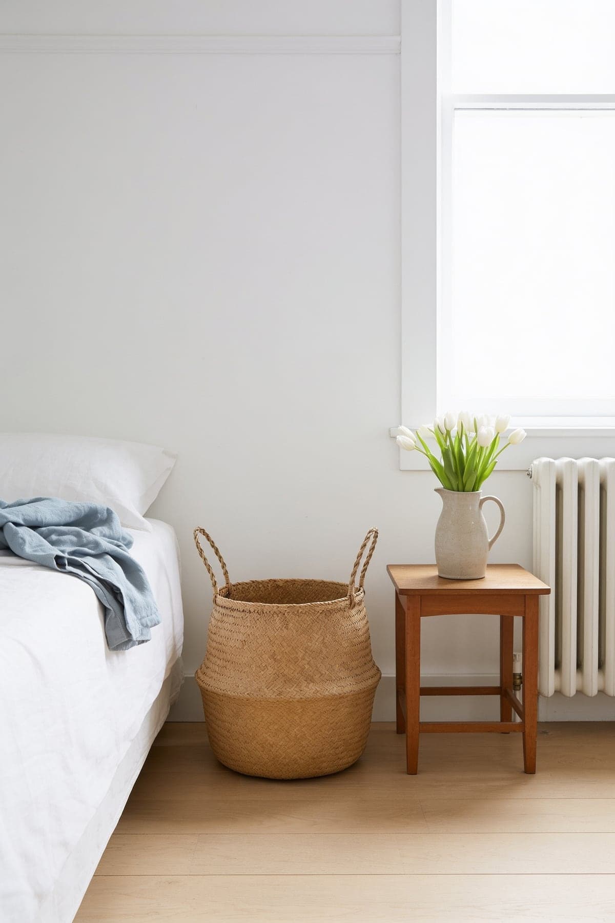 The seagrass belly basket beside a bed and a stool with tulips in a bright white bedroom