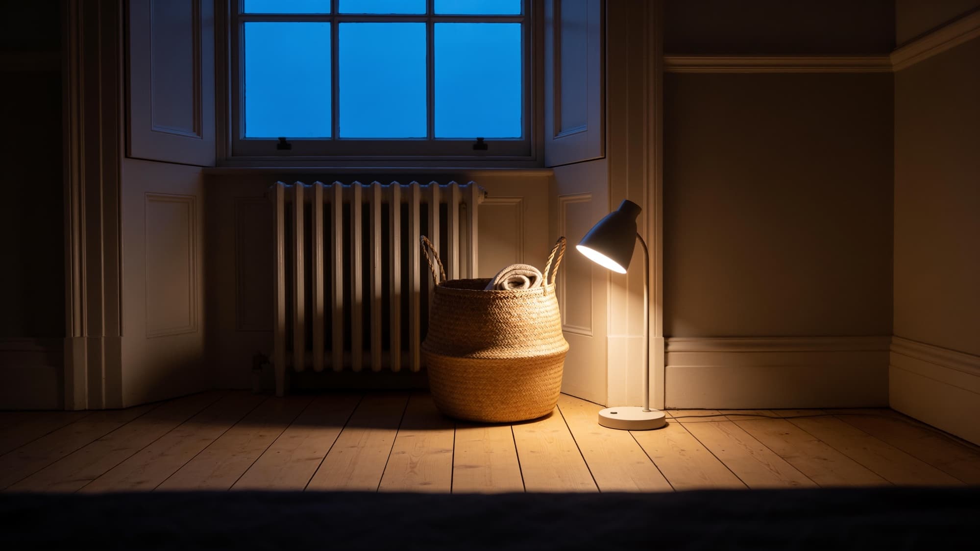 The seagrass basket holding a wool blanket on bare boards by a radiator at dusk