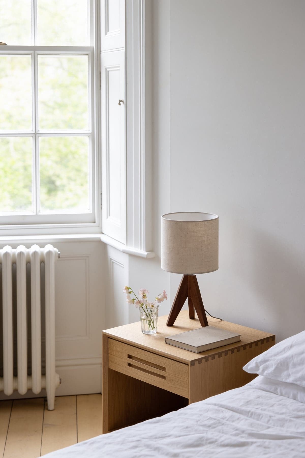 The wooden tripod lamp with its linen shade on an oak bedside table by a sash window
