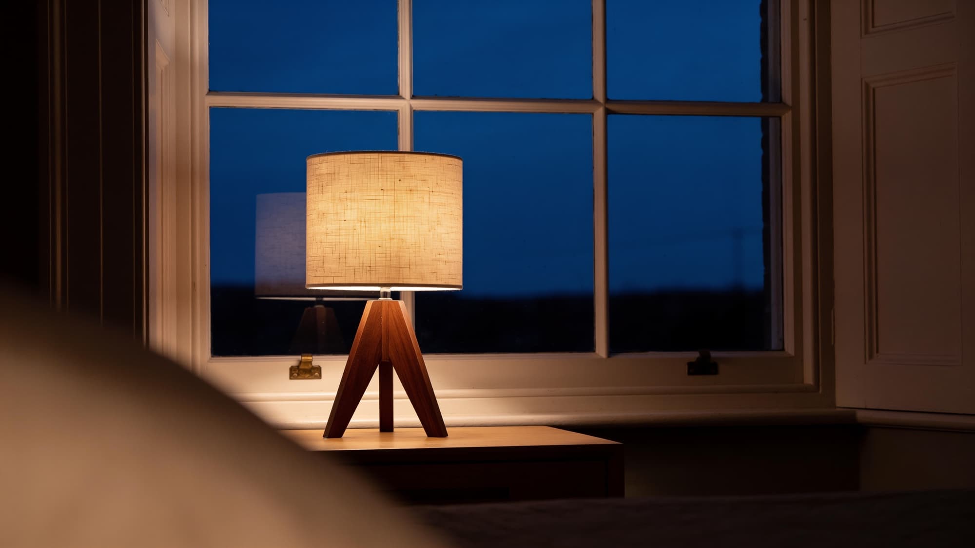 The tripod lamp glowing on an oak bedside table by a sash window at dusk