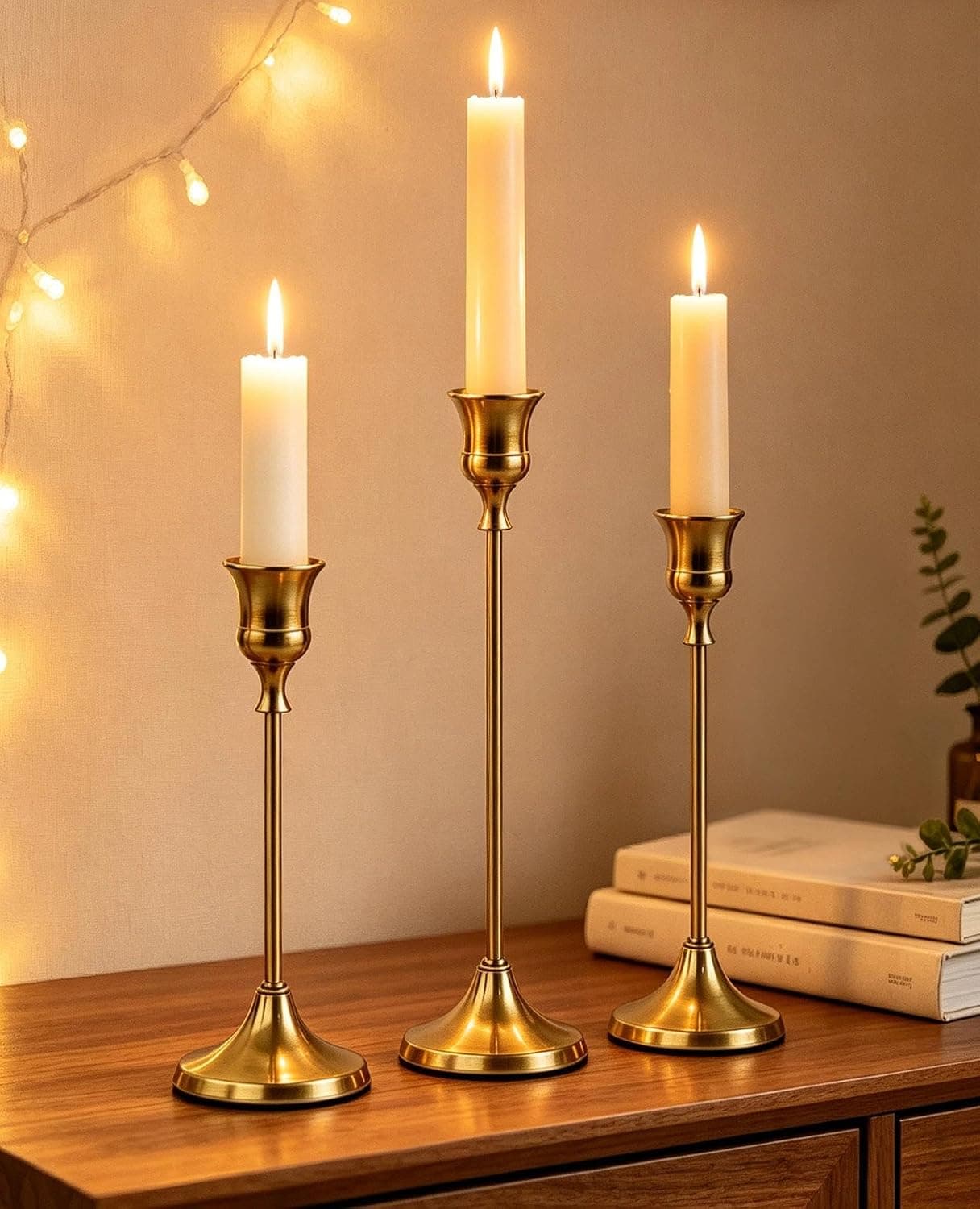 Three slim brushed gold candlesticks of different heights holding lit cream tapers on a wooden sideboard