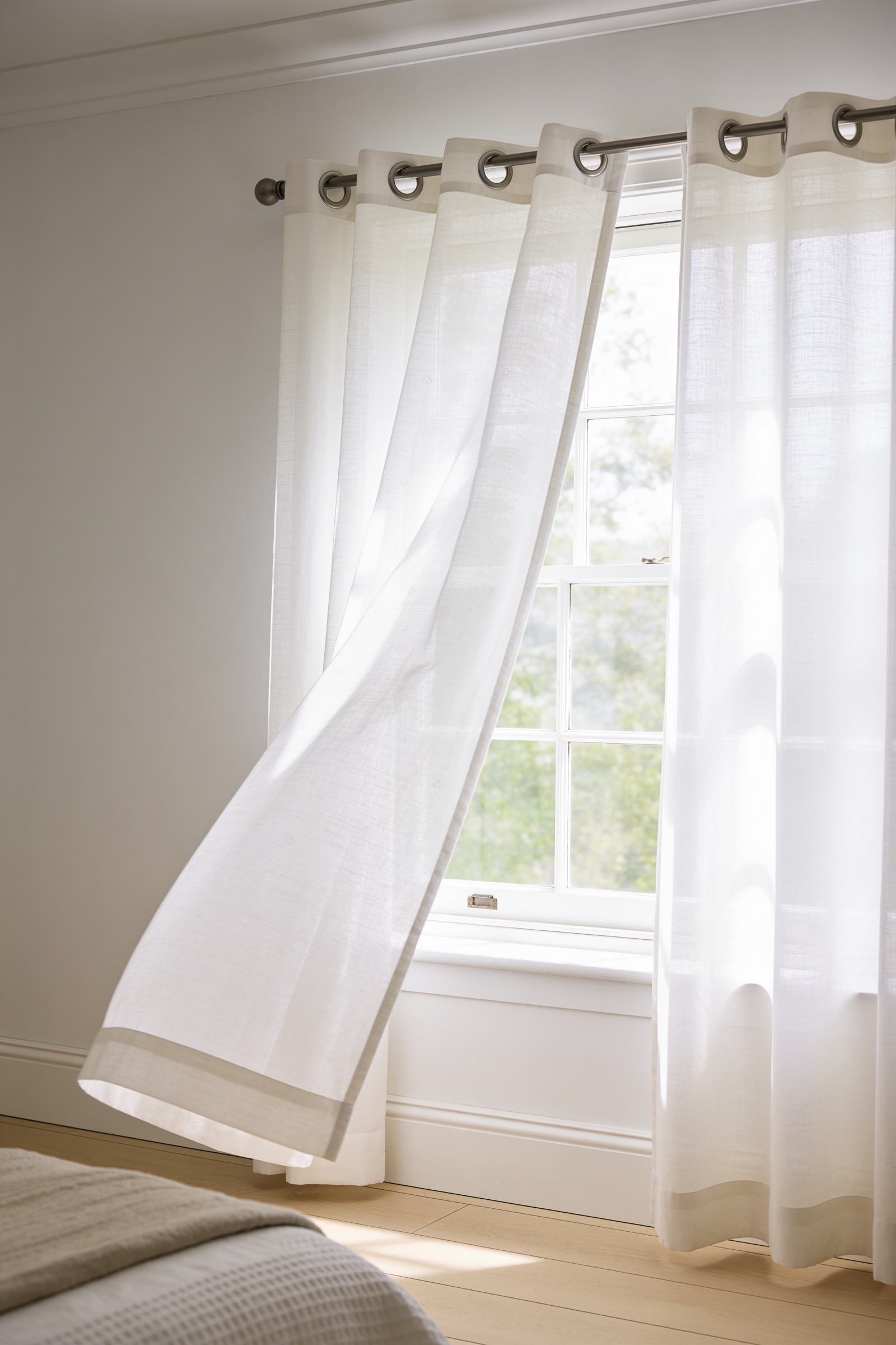 A pair of white woven linen-look voile eyelet curtains diffusing soft summer daylight at a bedroom window