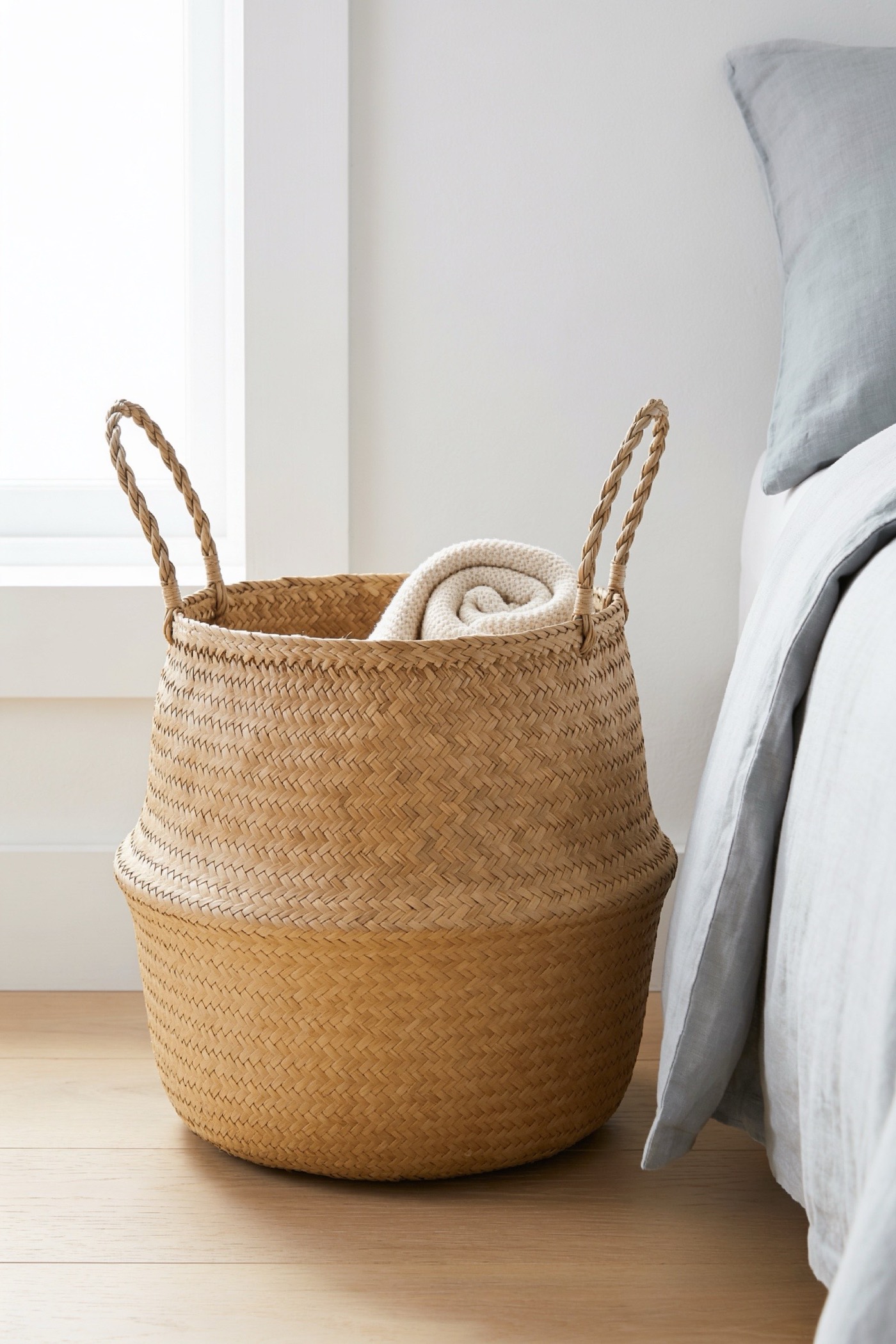 Woven natural seagrass storage basket with a handle and black jute trim beside a bed